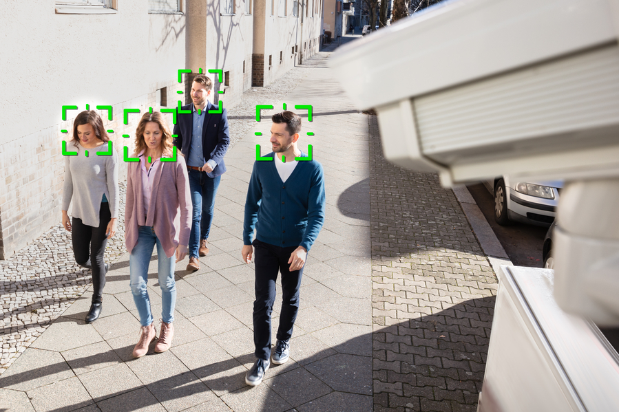A group of four people walking down a city sidewalk is being monitored by a surveillance camera, with green facial recognition boxes digitally overlaid on their faces.
