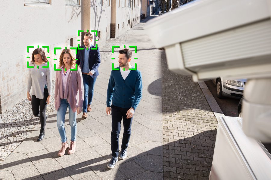 A group of people walking on a city sidewalk being tracked by an AI security camera with green facial recognition boxes highlighting their faces.