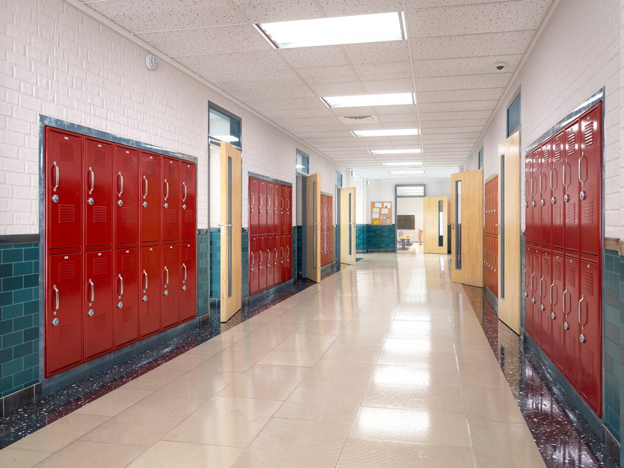 An empty school hallway with bright lighting, red lockers, and classroom doors open along both sides.