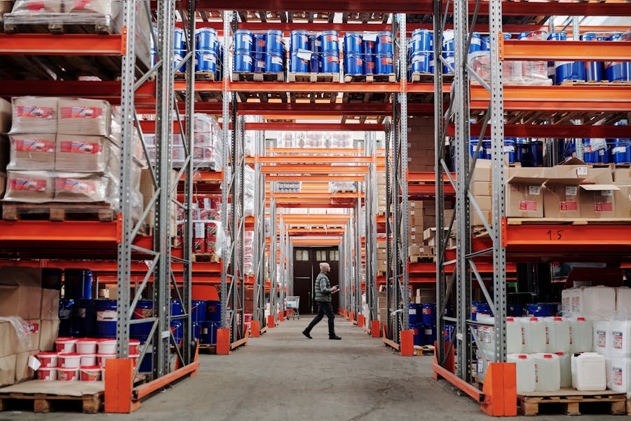 A worker walking through a large warehouse aisle lined with tall shelves filled with pallets, boxes, and industrial containers.