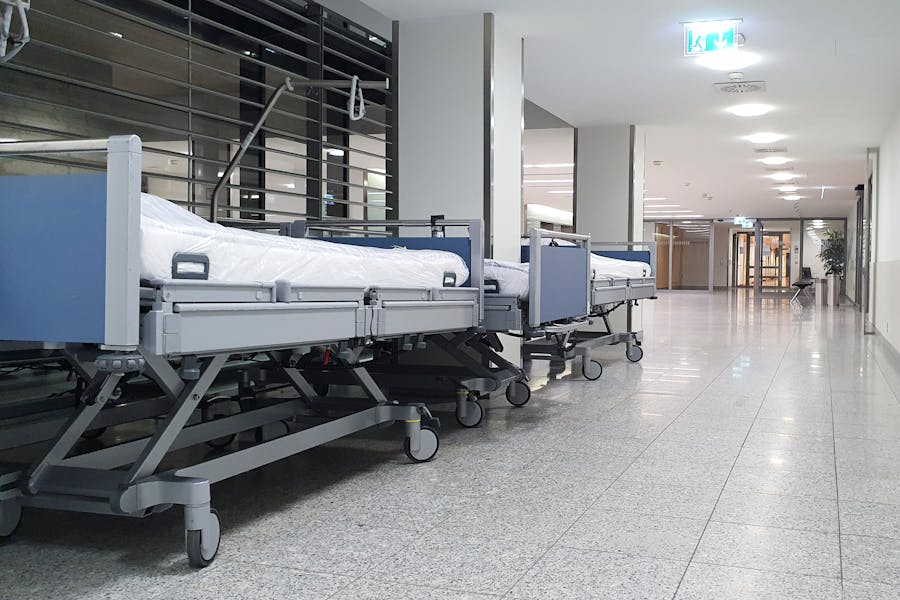 Empty hospital beds lined up in a bright, modern, and sterile medical facility hallway with polished tile floors.